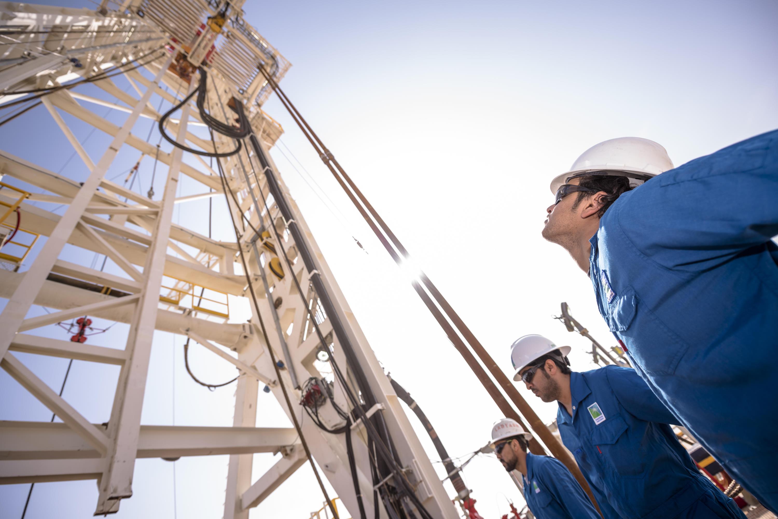 Saudi Aramco workers in hard hats look up at oil rig